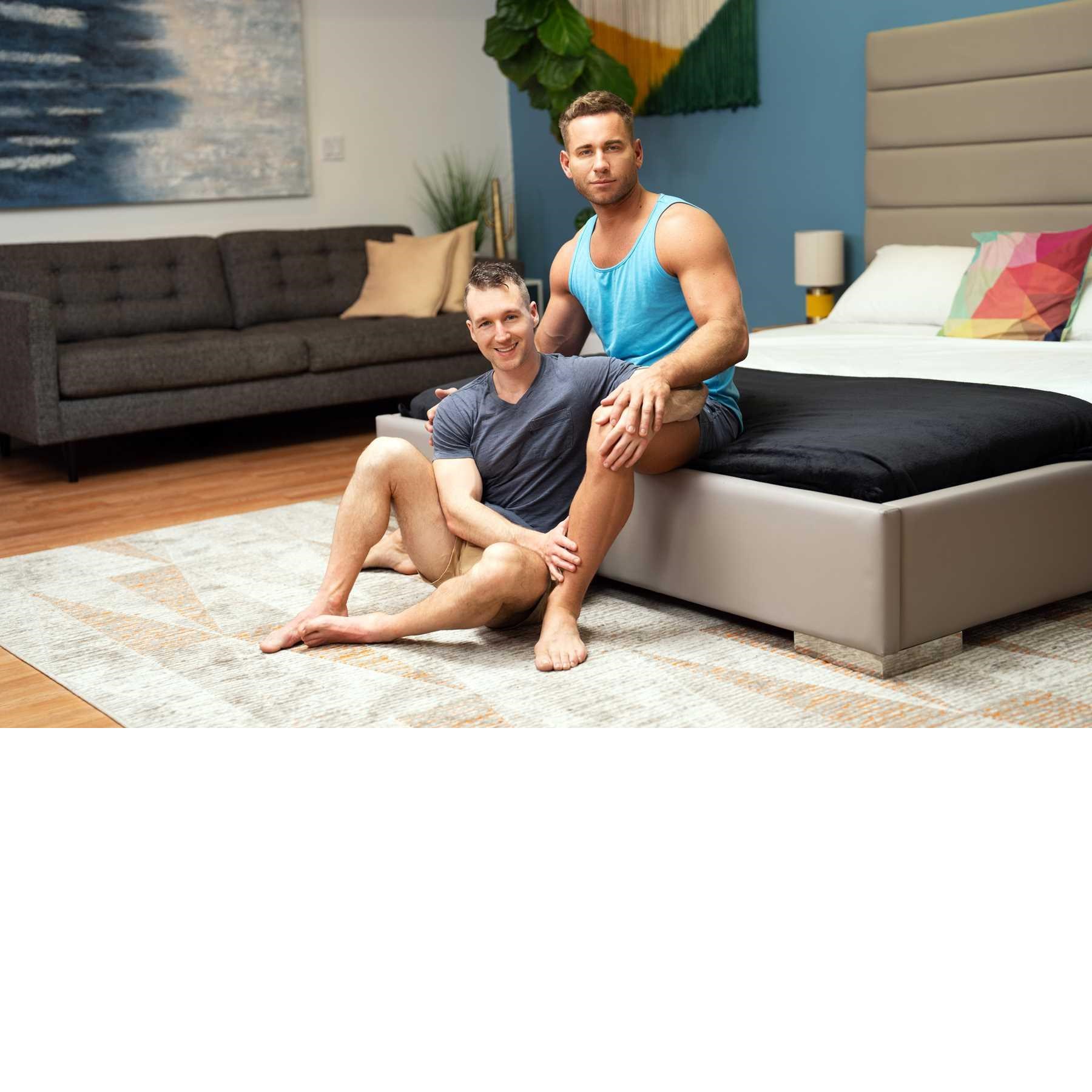 Two clothed males posed seated in bed room